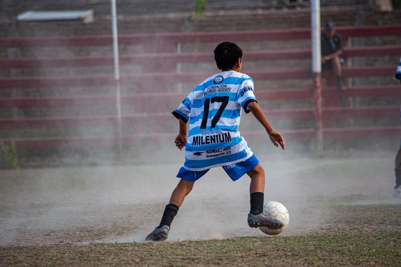A young soccer player wearing number 17 dribbles the ball on a dusty outdoor field.
